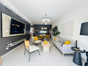 A modern living room with a flat-screen TV, a sofa, a dining area, and a kitchen in the background. - 2-bedroom apartment in fantastic Punta Cana with AC and gym (Punta Cana)