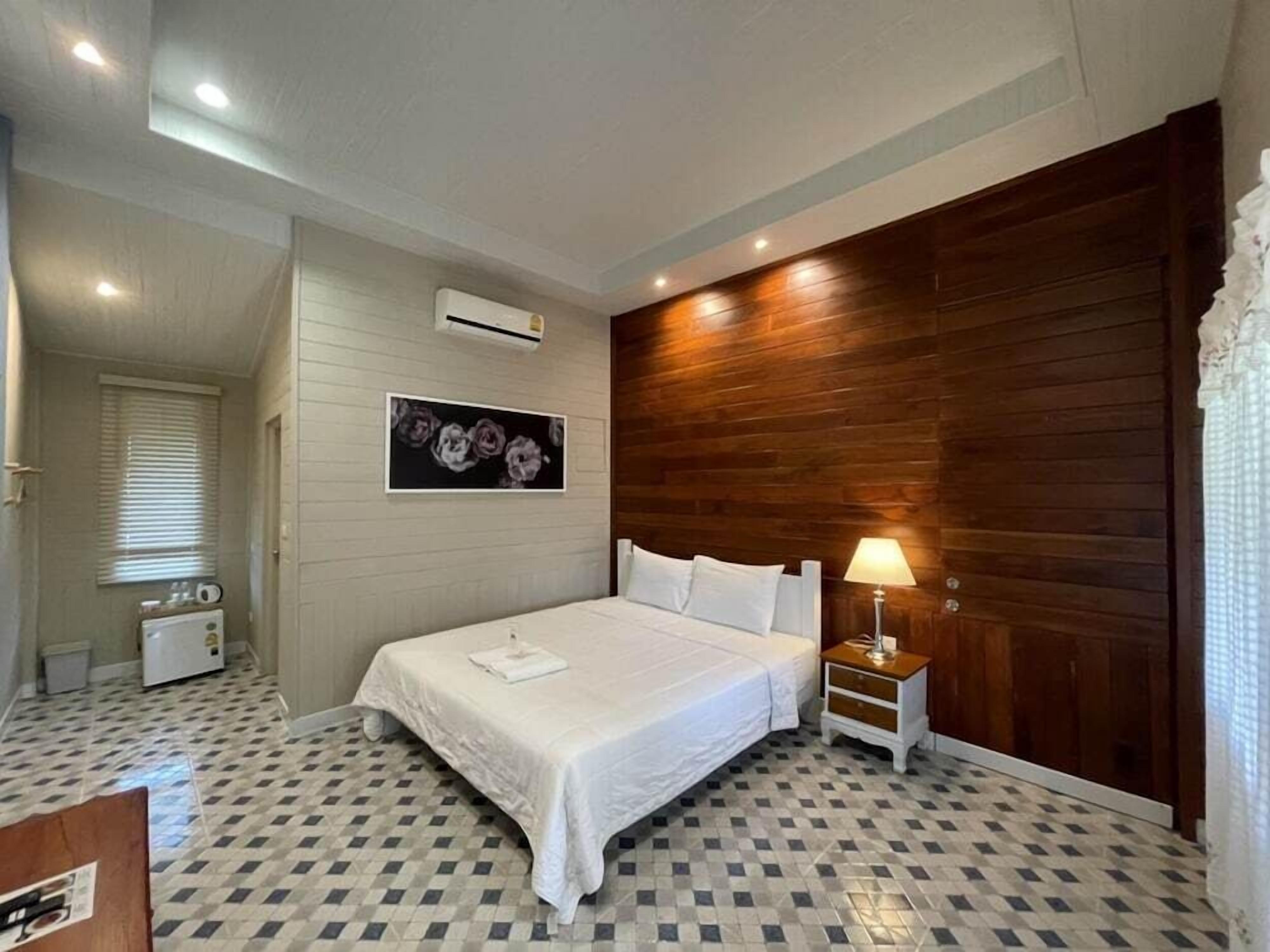 A bedroom with a checkered floor, a bed with white linens, a nightstand with a lamp, a wall-mounted air conditioner, and a framed floral artwork.