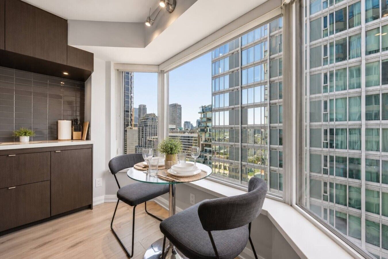 A modern kitchen with dark cabinetry, a round glass table, two chairs, and a view of a cityscape through large windows.
