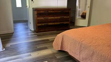A bedroom with a wooden dresser, a bed with a textured blanket, a flat-screen TV mounted above the dresser, and a hallway leading to another room.