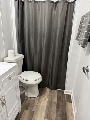 Un baño con un inodoro, un rollo de papel higiénico, un armario blanco y una cortina de ducha gris.
