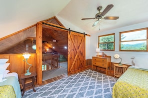 A bedroom with a lofted sleeping area, wooden walls, a ceiling fan, a bed, a dresser, a nightstand, and a view of the outdoors.