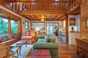 A cozy cabin interior with a wooden ceiling, a green sofa, a wicker chair, and a dining table.