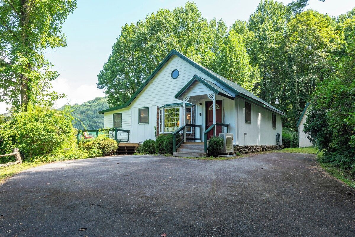 A single-story house with a front porch, a driveway, and a surrounding wooded area.