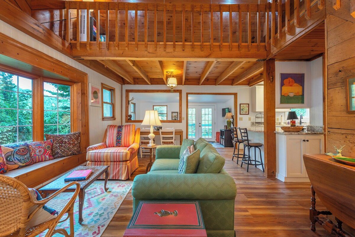 A spacious living room with a high ceiling, wooden beams, a green sofa, and a dining area with a wooden table and chairs.