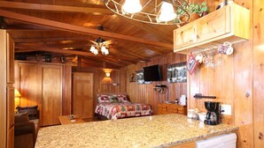 A wooden cabin interior with a kitchen area, a bed with a patterned blanket, and a television mounted on the wall. - Moose Haven Cabin, full kitchen, cover carport, Restmore Inn, Hill City (Hill City)