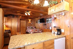 A wooden cabin interior with a kitchen area, a bed with a patterned blanket, and a television mounted on the wall. - Moose Haven Cabin, full kitchen, cover carport, Restmore Inn, Hill City (Hill City)