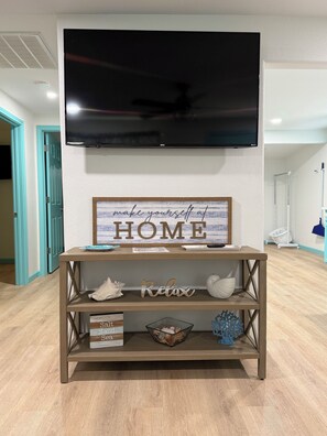 Ein Wohnzimmer mit an der Wand montiertem Fernseher, einem Holzkonsolentisch mit Dekoration und einem Schild mit der Aufschrift 'fühlt Euch wie ZUHAUSE'.