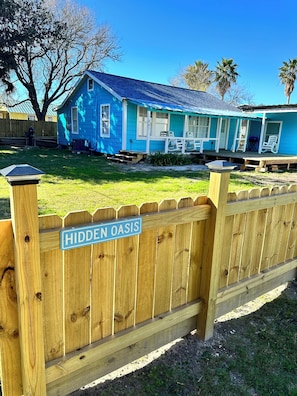 Ein blaues Haus mit Veranda und Holzzaun, an dem ein Schild mit der Aufschrift 'Hidden Oasis' steht.