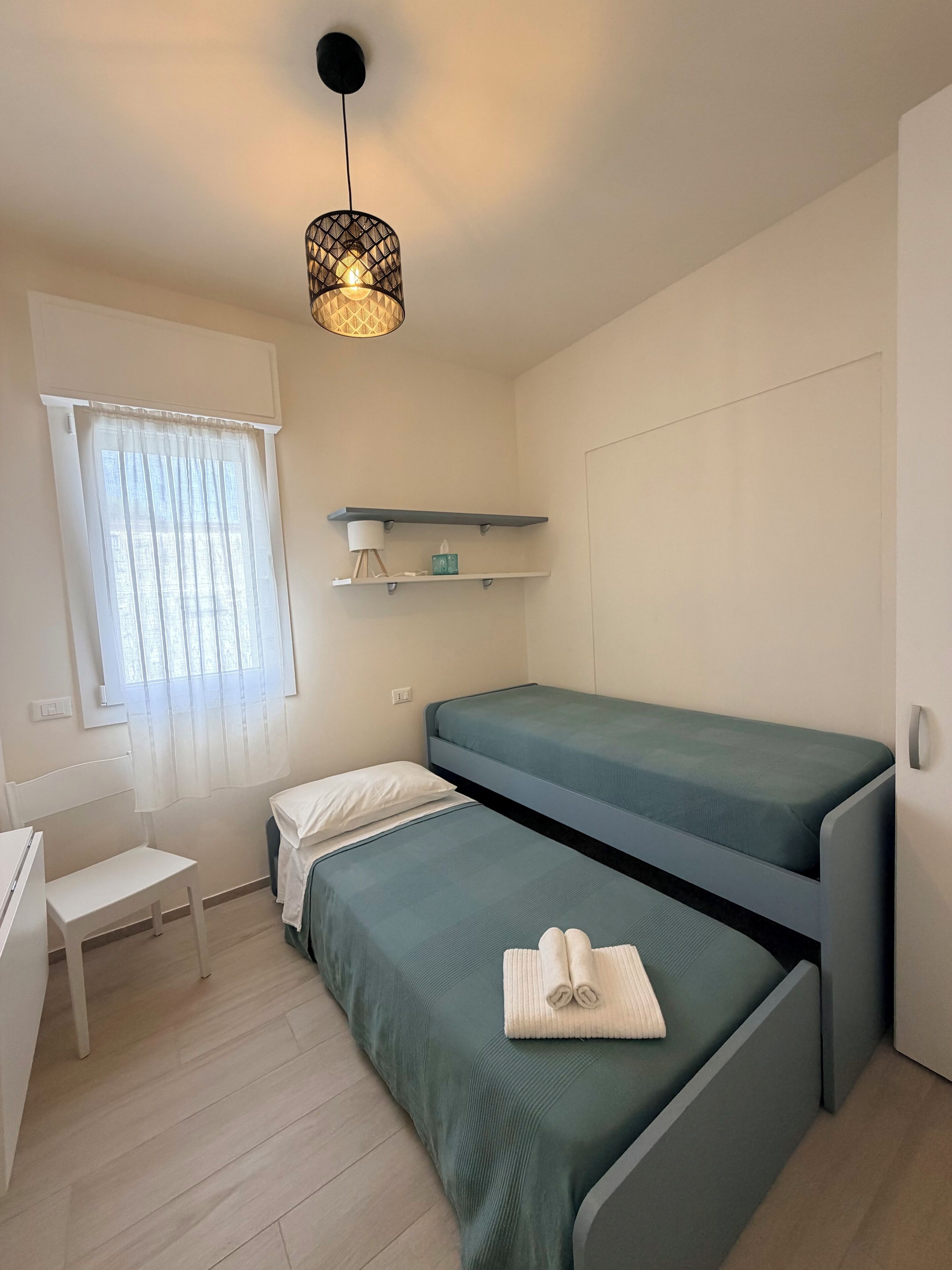 A small, modern room with two bunk beds, a desk, a chair, a hanging light fixture, and a window with sheer curtains.