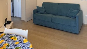 A living room with a blue sofa, a dining table with a patterned tablecloth, a TV mounted on the wall, and an air conditioning unit. - Modern Sea Apartment (Punta Marina)