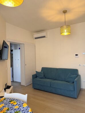Ein Wohnzimmer mit einem blauen Sofa, einem Esstisch mit gemustertem Tischläufer, einem an der Wand befestigten Fernseher und einer Klimaanlage.