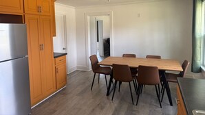 Cocina moderna con armarios de madera, refrigerador de acero inoxidable, mesa de comedor de madera con sillas y vista a un pasillo.