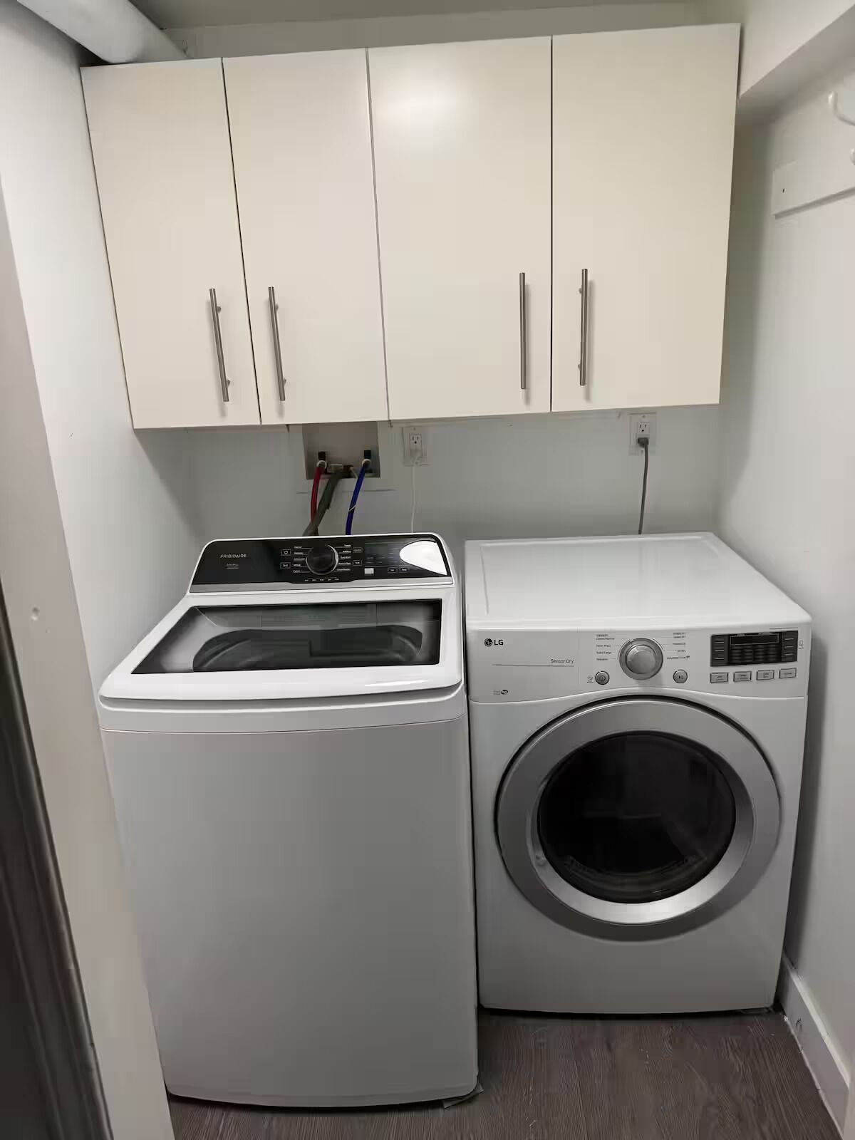Uma lavanderia compacta com máquina de lavar e secadora de abertura frontal, armários brancos e tomadas elétricas visíveis.