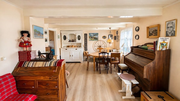 A cozy living room with a wooden sofa, a dining table with chairs, a piano, and a kitchen area with cabinets and a stove.