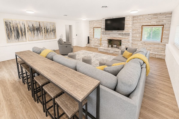 Un salon moderne avec un canapé d’angle, une table à manger en bois avec des bancs, un téléviseur à écran plat fixé à un mur en pierre et une cheminée.