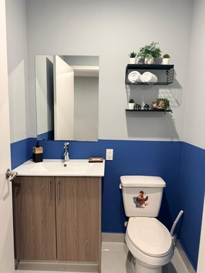A bathroom with a white sink, wooden cabinets, a toilet with a rooster decal, a mirror, and a shelf with plants and decorative items. - Spacious Florida Keys Escape. Family Friendly (Florida City)