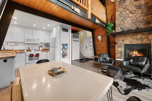 A modern kitchen with white cabinets, a dining table, and a fireplace with a stone wall backdrop. - Boutique Cabin, Across From Zoo, Mountain Views (Big Bear Lake)