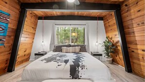 Ein Schlafzimmer mit einem großen Bett, einem Fenster mit Blick auf Bäume, Holzwänden und einem Deckenventilator.