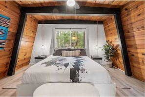 A bedroom with a large bed, a window with a view of trees, wooden walls, and a ceiling fan. - Boutique Cabin, Across From Zoo, Mountain Views (Big Bear Lake)