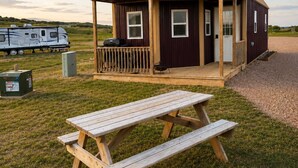 A small, single-story building with a porch, a picnic table, and a trailer in the background.