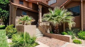 A two-story house with a brick exterior, a front porch, and a landscaped garden with palm trees. - Sports Village Condo | Pools, Courts & Trails (St. George)