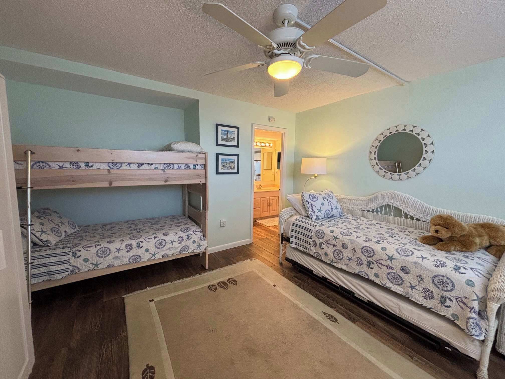 A room with bunk beds, a sofa, a ceiling fan, a mirror, and a door leading to a bathroom.