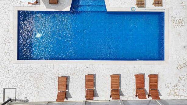 Ein rechteckiger Swimmingpool mit weiß gefliestem Sonnendeck, Holzliegen und einem weißen Sonnenschirm.