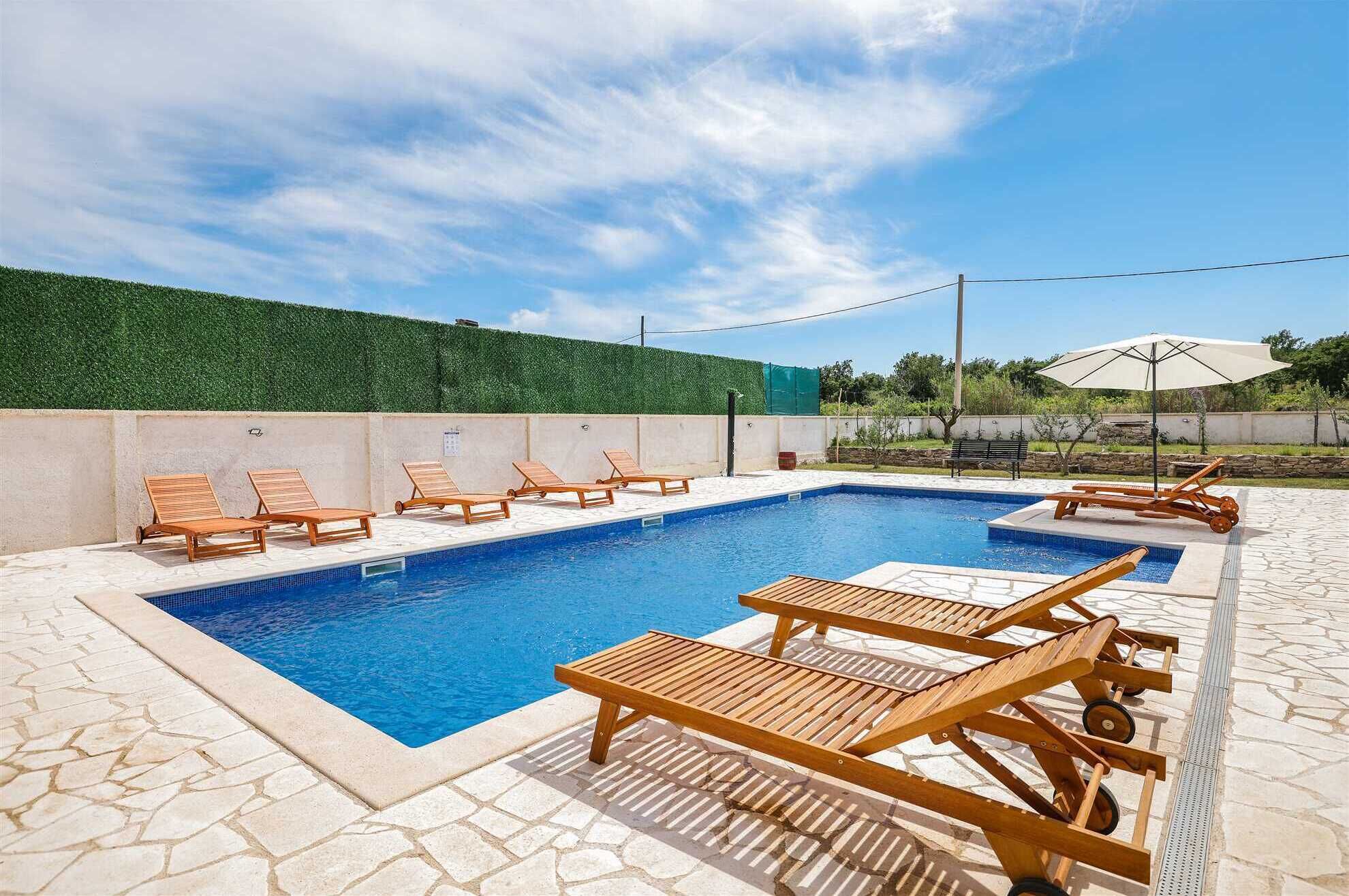 En swimmingpool med träloungestolar, ett däck med stenplattor och en grön häck.