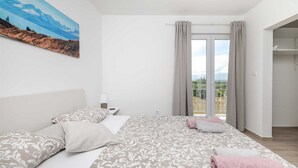 Ein Schlafzimmer mit Bett, Kissen, einem Nachttisch, einem Gemälde und einem Balkon mit Vorhängen.