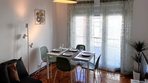 Zona pranzo con tavolo imbandito per quattro persone, lampada da terra, pianta in vaso e tende a motivi decorativi.