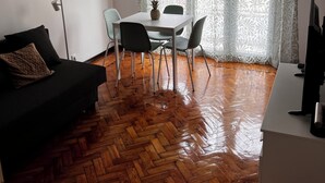 Una stanza con pavimento in legno a spina di pesce, un tavolo da pranzo con sedie, un divano nero, una lampada da terra e un televisore.