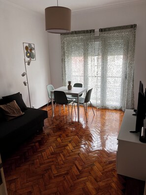 Ein Zimmer mit Fischgrätparkett, einem Esstisch mit Stühlen, einer schwarzen Couch, einer Stehlampe und einem Fernseher.