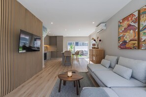 Een moderne woonkamer met een flatscreen televisie, een grijze bank, een houten accentmuur en een eethoek met stoelen en een tafel.