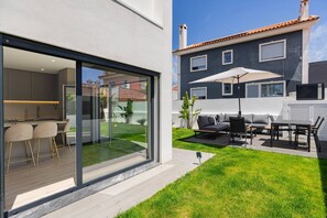 Een modern huis met een glazen deur naar een keuken, een terras met tuinmeubilair, en uitzicht op een naburig huis.