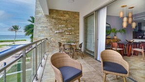 A balcony with wicker chairs, a stone wall with fish decorations, a view of the beach, and a kitchen area with wooden furniture. - Oceanfront Condo w/ Pool & Balcony, Costa (Punta Cana)