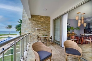 A balcony with wicker chairs, a stone wall with fish decorations, a view of the beach, and a kitchen area with wooden furniture. - Oceanfront Superior Condo with Pool in Punta Cana (Punta Cana)