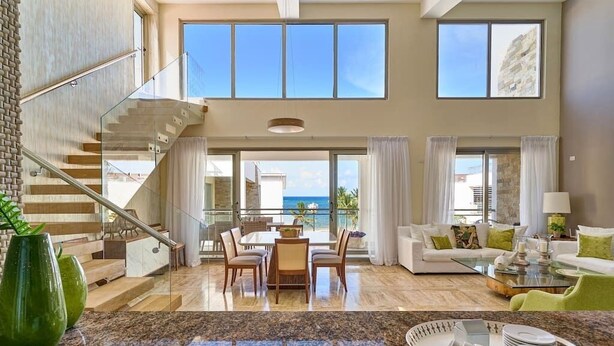 A spacious living area with a dining set, a staircase, and large windows offering a view of the sea. - Oceanfront Penthouse w/Rooftop, Pool, Punta Cana (Punta Cana)