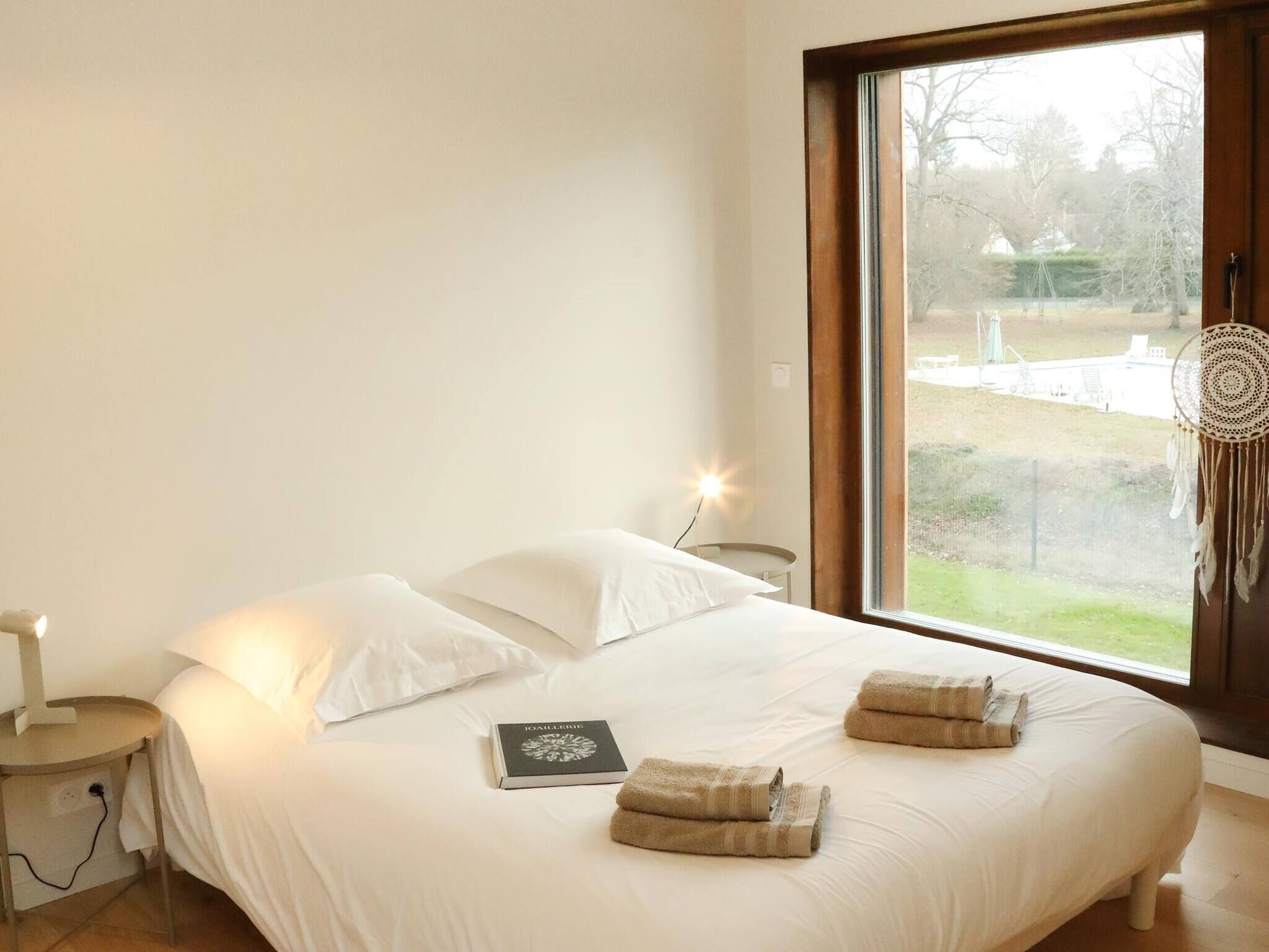 Une chambre avec une grande fenêtre, un lit recouvert de draps blancs, une table de chevet, une lampe et un livre.
