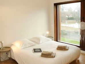 Un dormitorio con un ventanal grande, una cama con ropa de cama blanca, una mesita de noche, una lámpara y un libro.