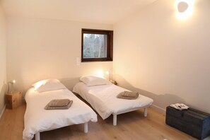 Una habitación con dos camas individuales, una ventana, una mesita de noche con lámpara y un pequeño cofre de madera.