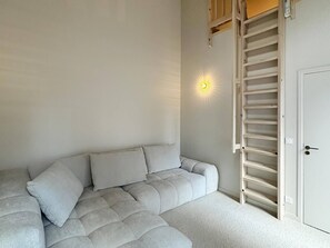 Una habitación con sofá modular, una lámpara de pared, una escalera de madera y una puerta.