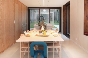 Un comedor moderno con una mesa blanca, una silla azul y vistas a un jardín a través de grandes ventanales.