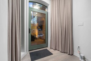 Een moderne entree met een glazen deur, beige gordijnen, een deurmat en een stopcontact op de muur.