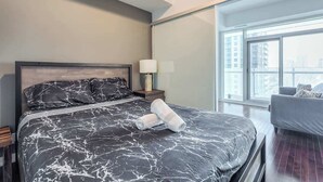 Quarto de hotel moderno com uma cama grande, uma mesinha de cabeceira com luminária, uma cadeira e uma janela ampla com vista para a cidade.