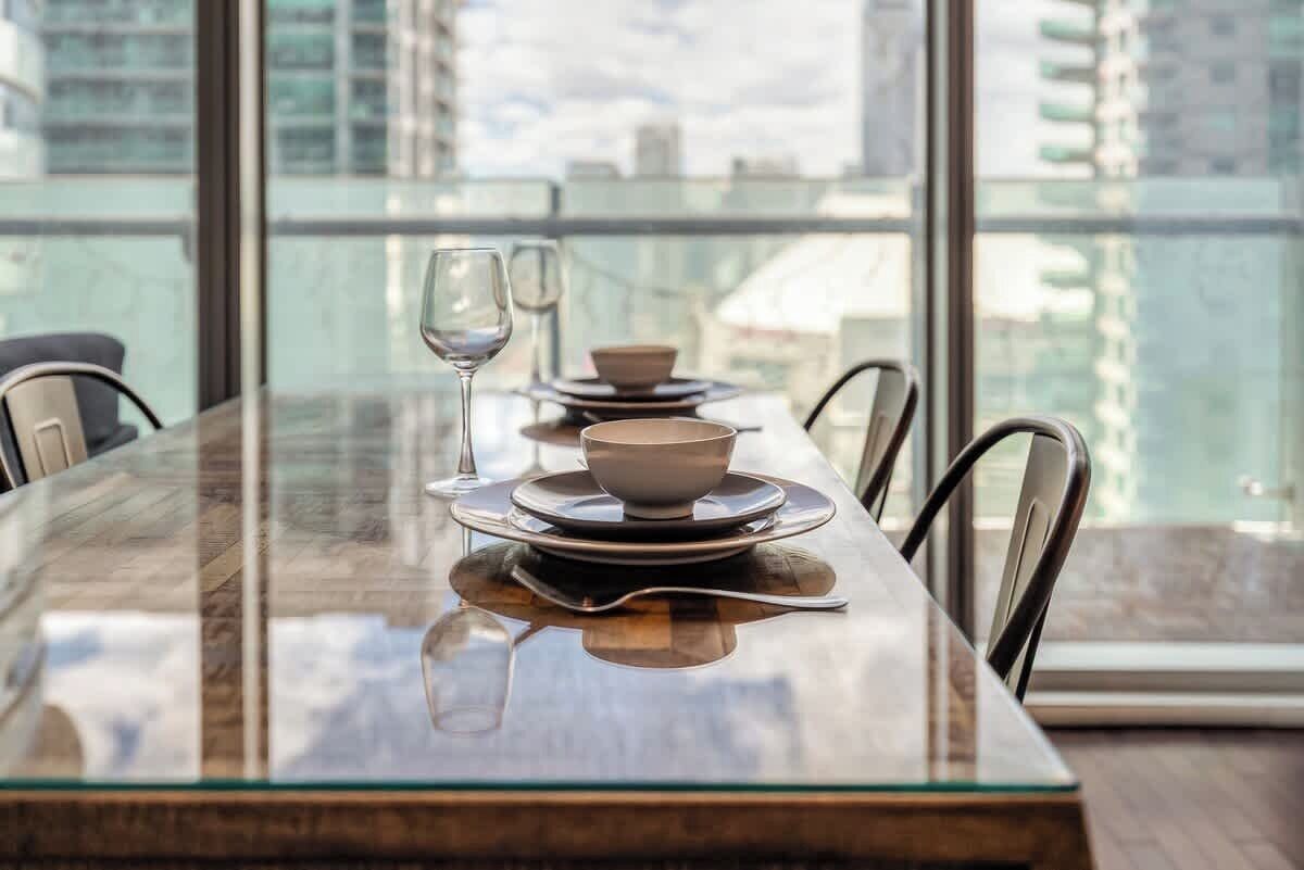 A dining table with plates, bowls, and wine glasses set for two, with a view of buildings through large windows.