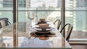 A dining table with plates, bowls, and wine glasses set for two, with a view of buildings through large windows.