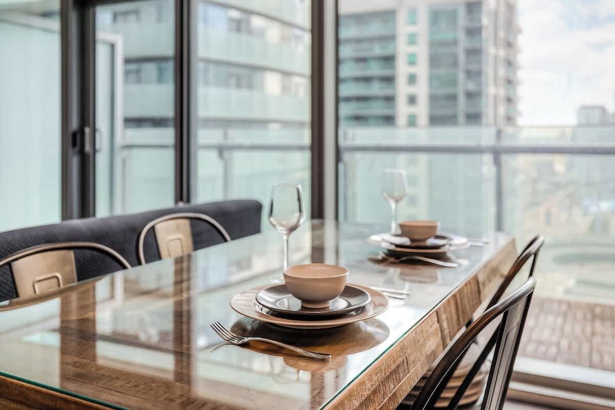 A dining table with a glass top, two chairs, and a view of a cityscape through large windows.