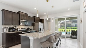 A modern kitchen with dark cabinets, a large island, bar stools, and a view of a landscaped outdoor area. - Chic Orlando Escape Game Room Theme Beds Pool (Clermont)