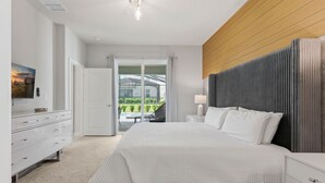 A modern bedroom with a large bed, a wooden headboard, a white dresser, and a view of a balcony with greenery. - Chic Orlando Escape Game Room Theme Beds Pool (Clermont)